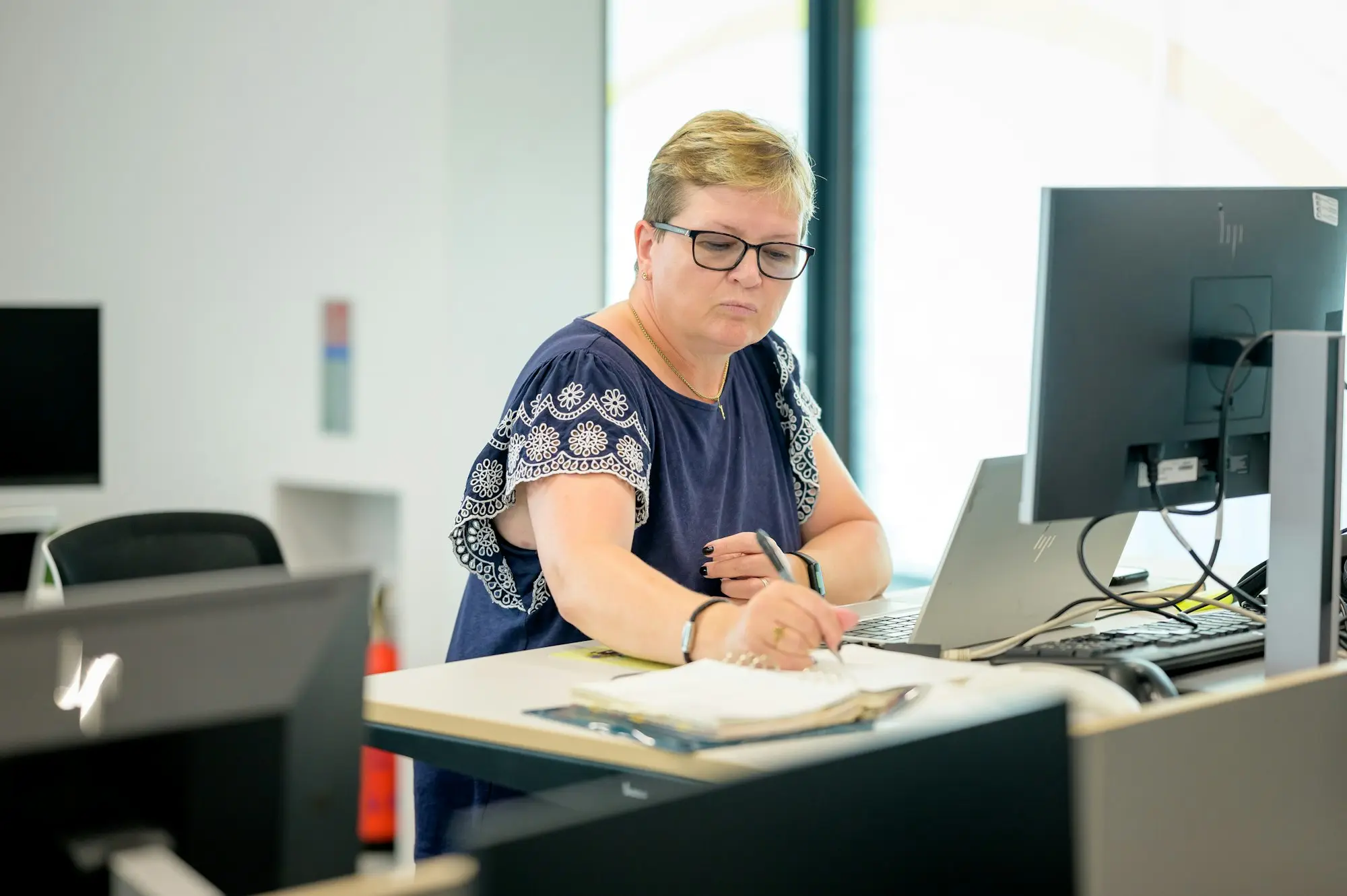 A woman at a small office desk with a monitor and notebook, looking up from her bookkeeping work.