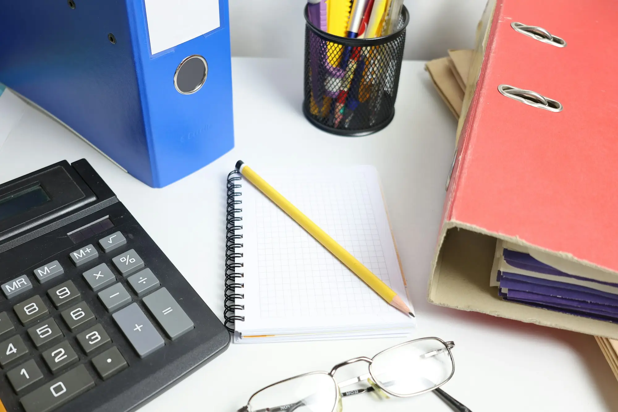 Desk with blue and pink folders, a calculator, notepad, pencil and glasses ready for end-of-year paperwork.