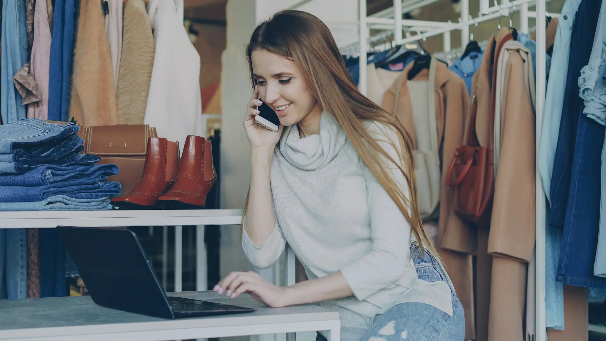 Shop owner on a mobile phone call at a retail counter with a laptop open.