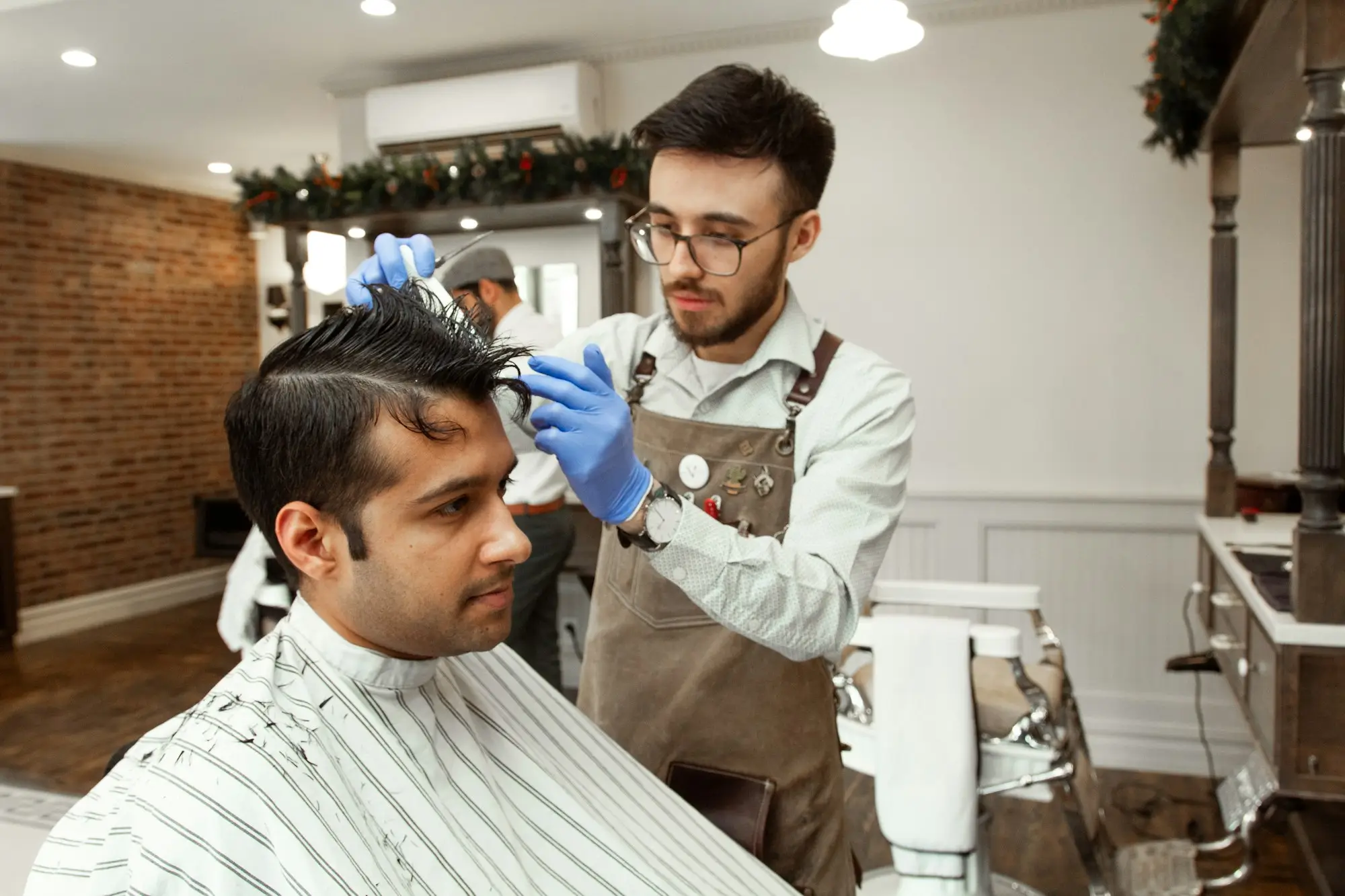 Barber employee in a work apron and gloves styling a client's hair in a small barbershop with Christmas garland on the walls
